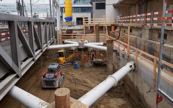 Des ouvriers et des engins de construction se trouven dans une fouille à côté de voies ferrées.
